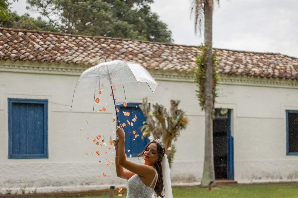 Noiva com sombrinha transparente diante da fachada caiada, sob chuva de pétalas