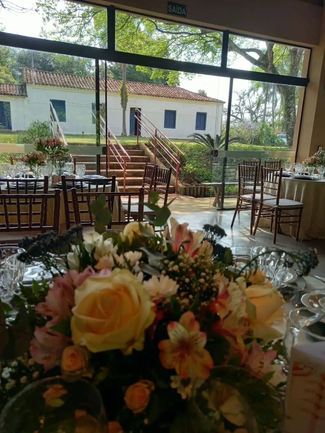 Vista de dentro do salão envidraçado para o gramado, com a casa do outro lado e as mesas postas em primeiro plano
